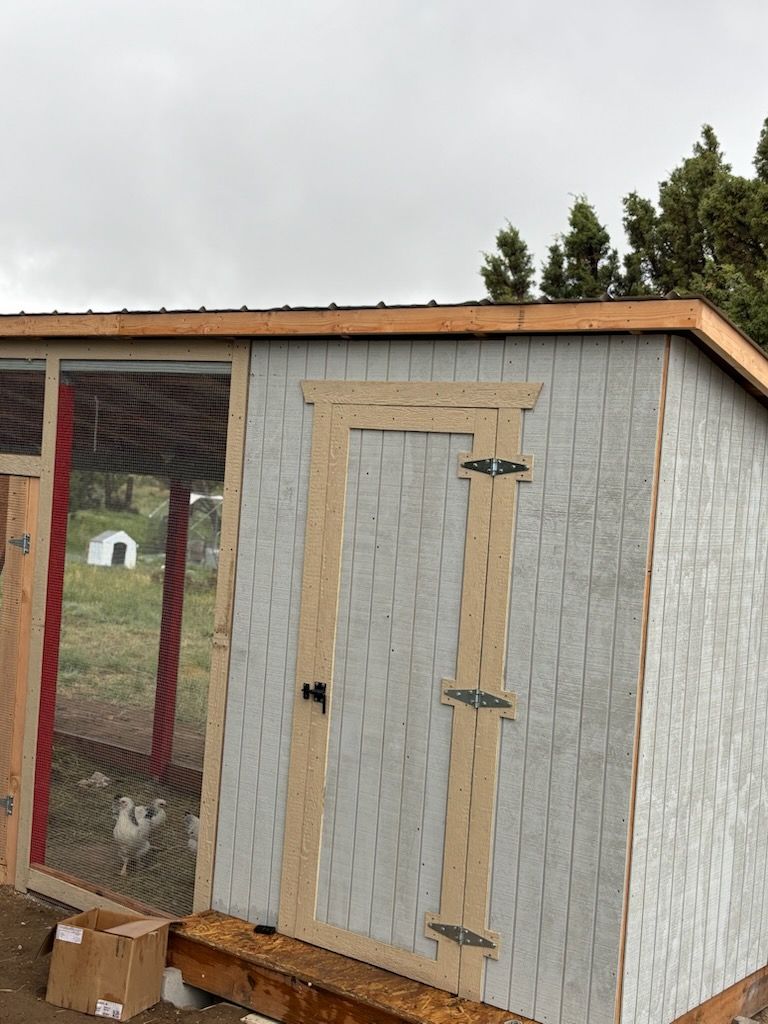 Small gray shed with tan roof and red-framed door beside a fenced area.