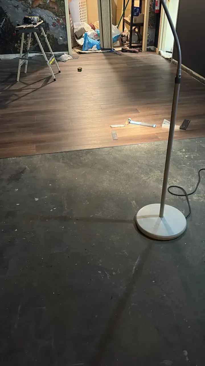 Dim room with a wooden floor panel under construction and a standing floor lamp in the foreground.