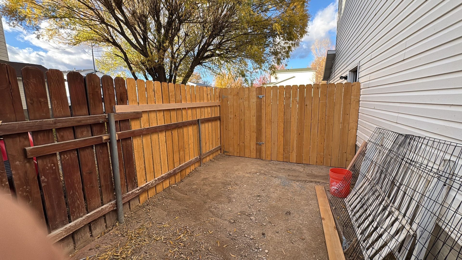 Small gravel backyard corner with wooden fence, tree, and house siding, with a red item near the wall