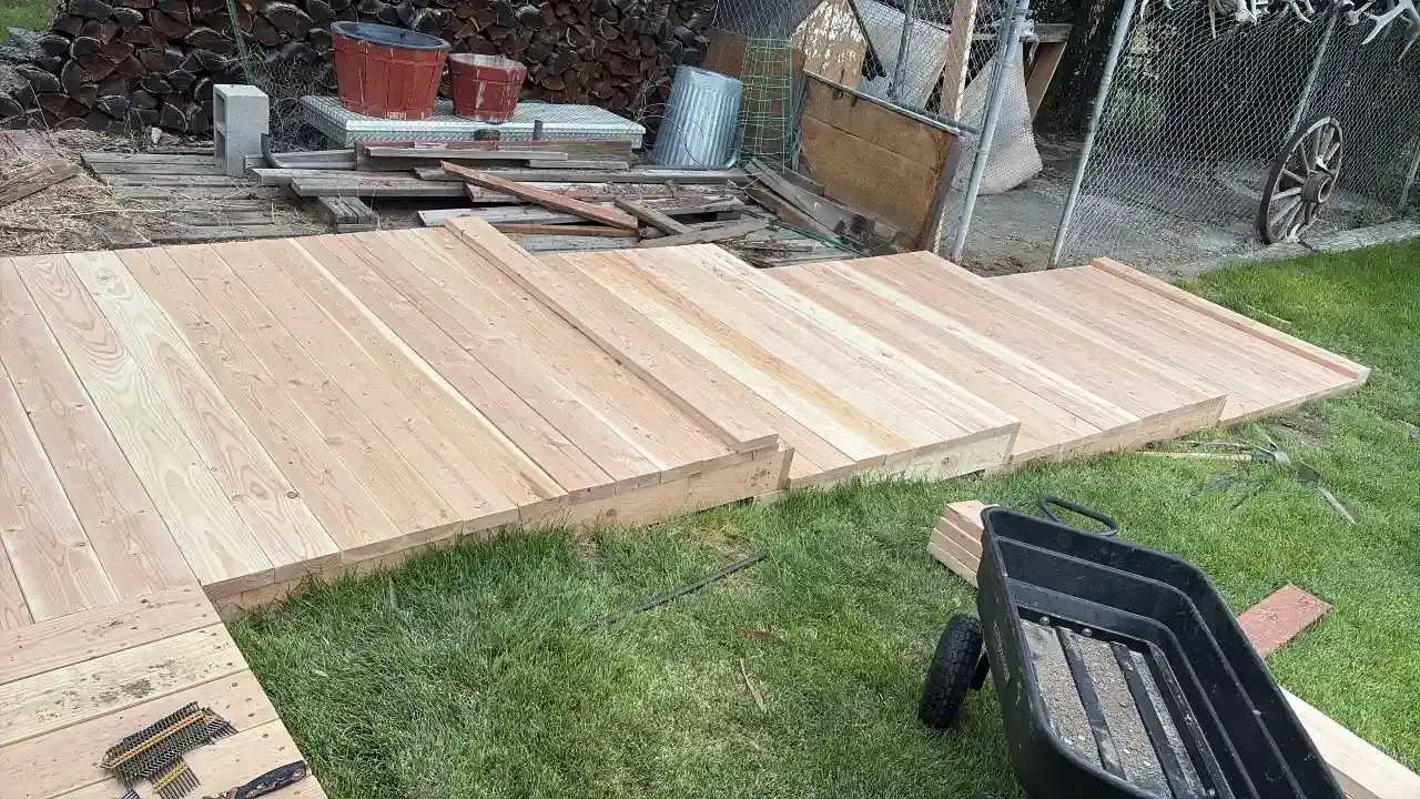 Wooden deck boards laid out on grass in a backyard beside a fence and stacked materials.