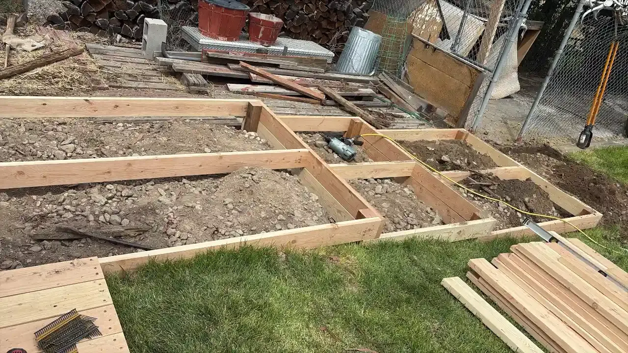 Wooden foundation framing laid out on dirt beside a patch of grass and stacked lumber.