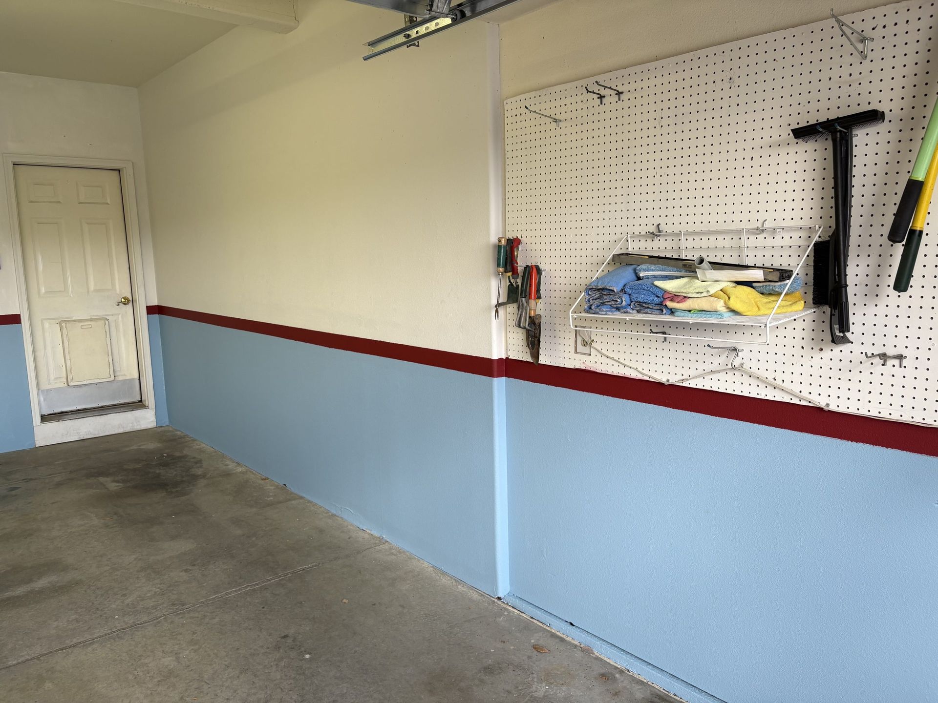 Empty garage with light blue walls, red trim, pegboard tools, and a white door.