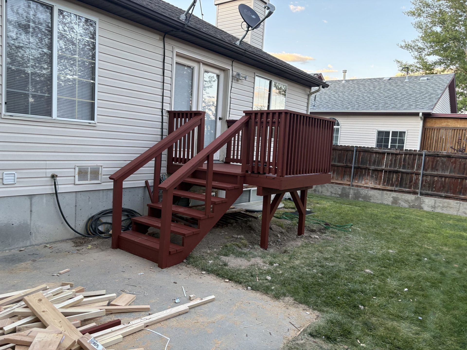 Backyard wooden deck stairs and railing beside a beige house, with lawn and lumber debris nearby