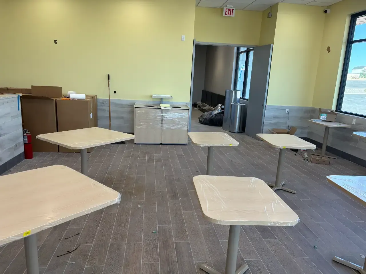 Empty cafeteria with small square tables, brown tile floor, yellow walls, and stainless steel counters.