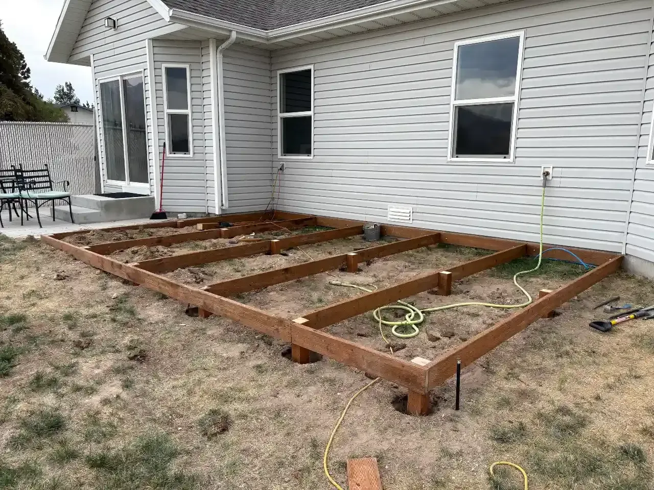 Wooden deck frame under construction beside a white house on a patchy lawn