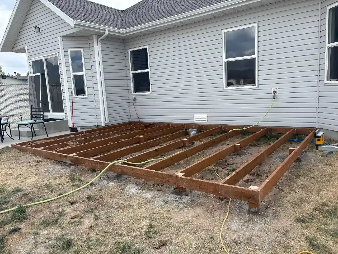 Backyard deck under construction beside a white house, with exposed wooden framing and dirt ground