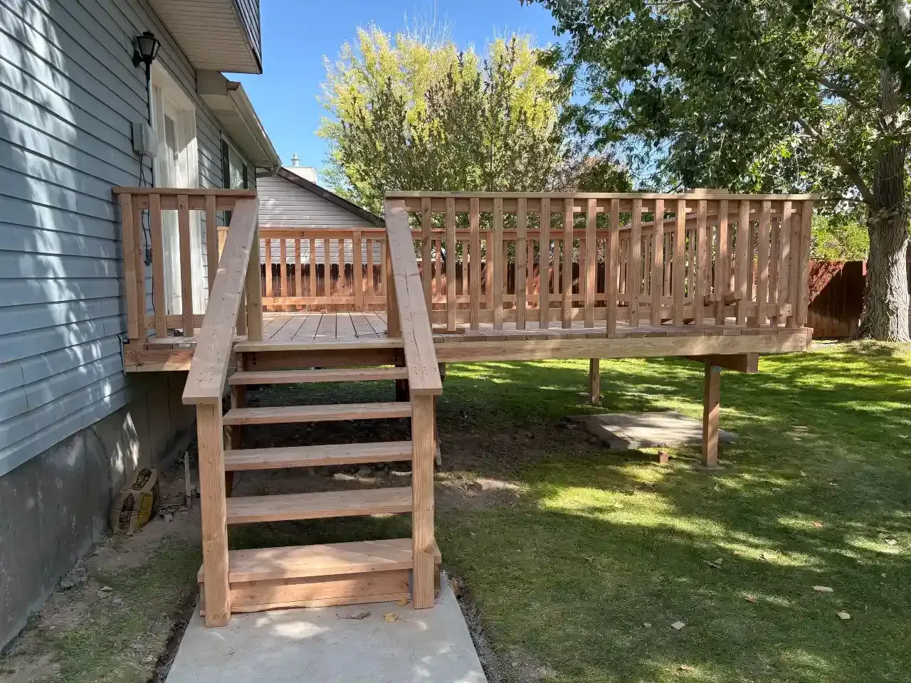 Wooden backyard deck with stairs beside a house, surrounded by grass and trees.