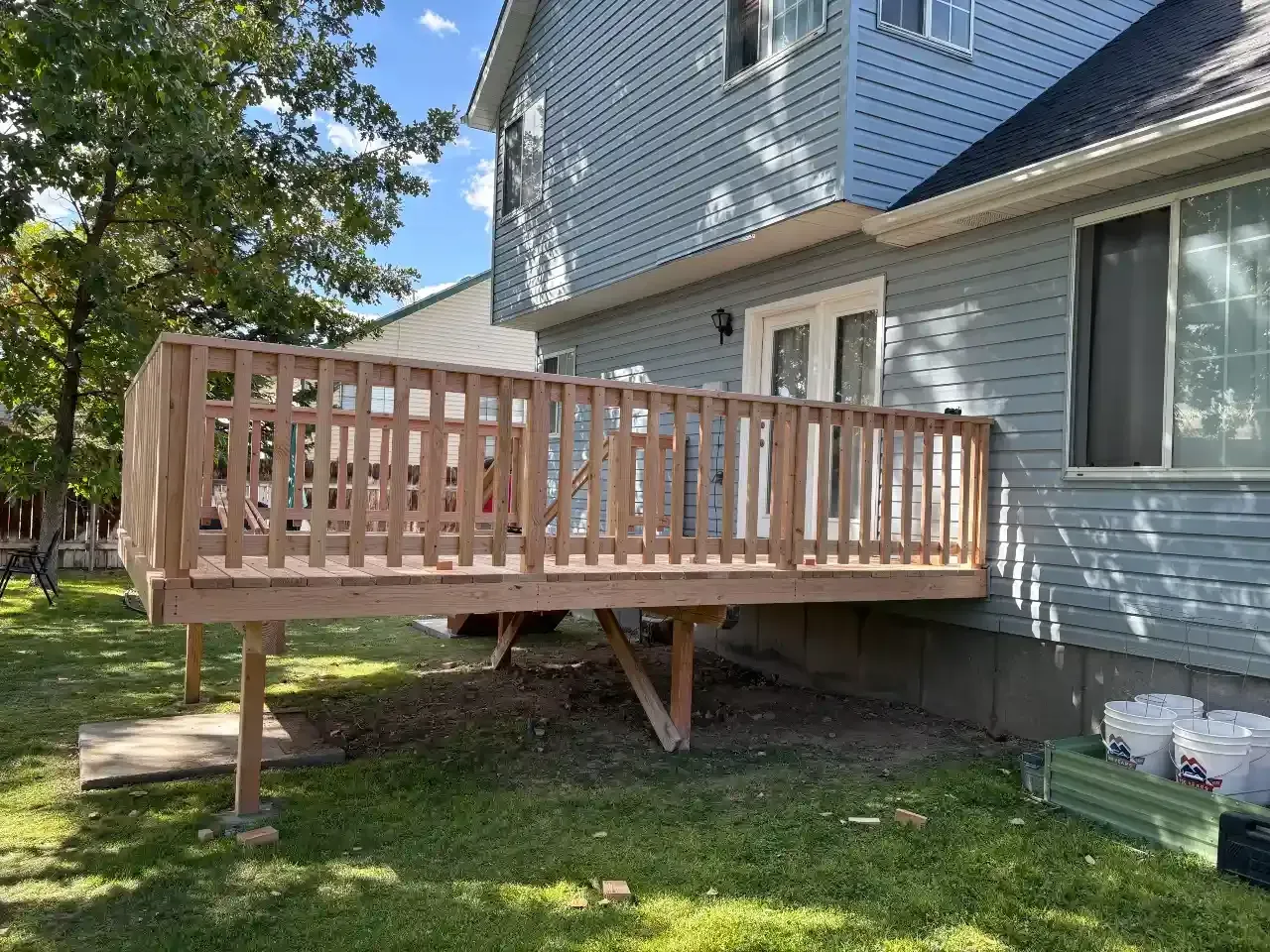Wooden deck addition beside a blue house, with support posts and railing on a grassy yard.