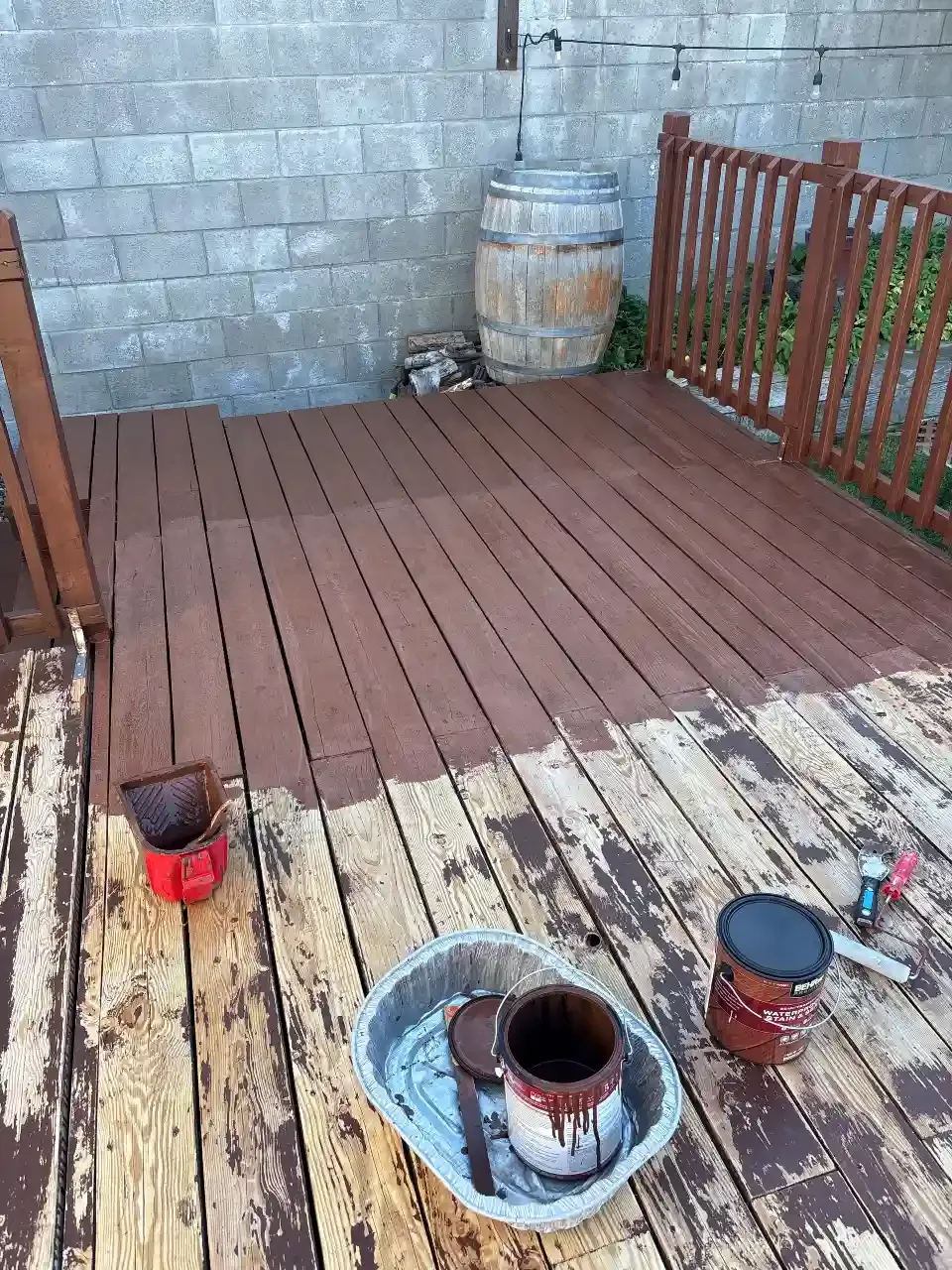 Small patio with a new brown wooden deck beside old boards, paint buckets, and a concrete wall.