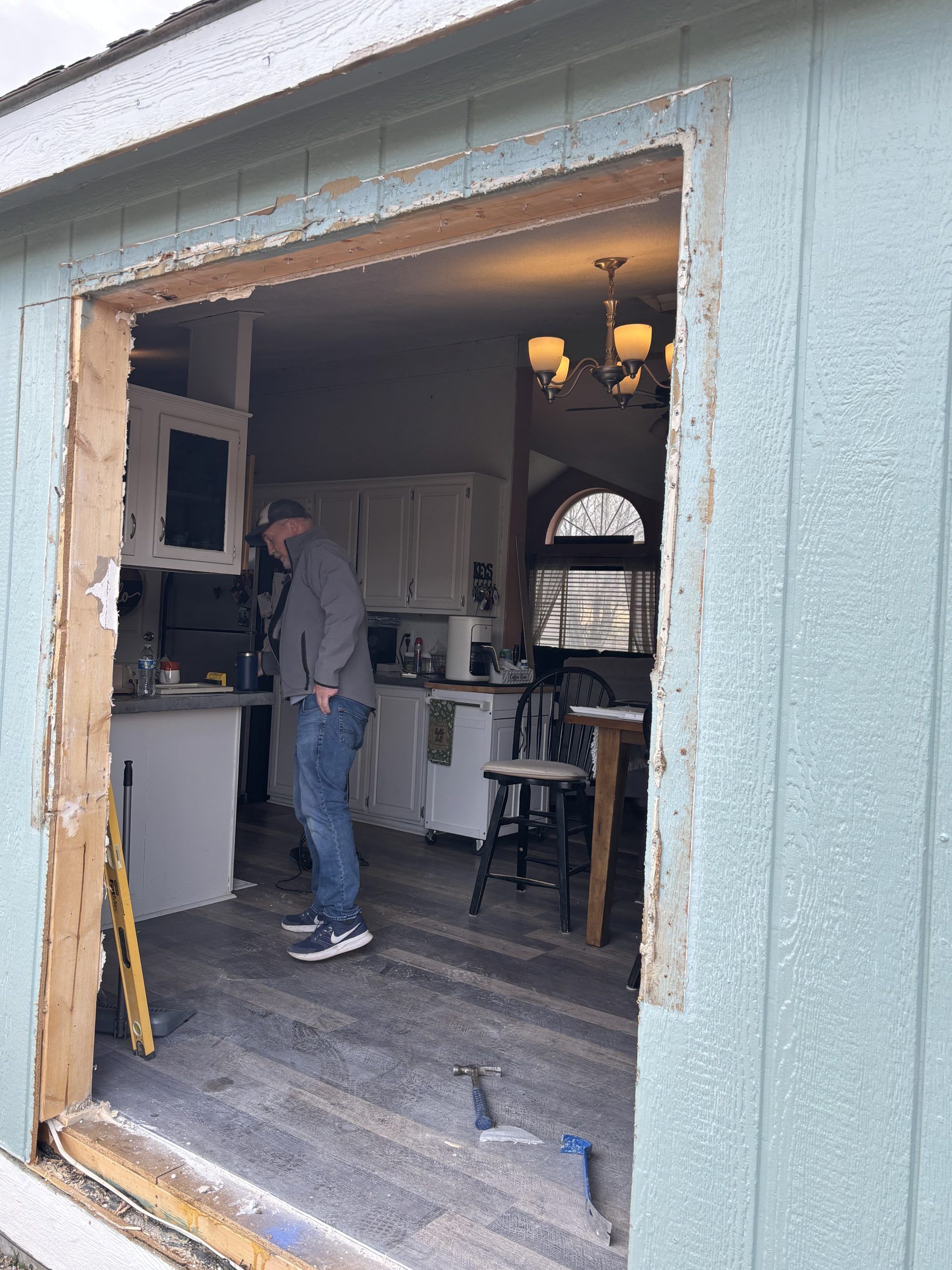Worker standing in a house doorway during a renovation, with exposed framing and tools inside.