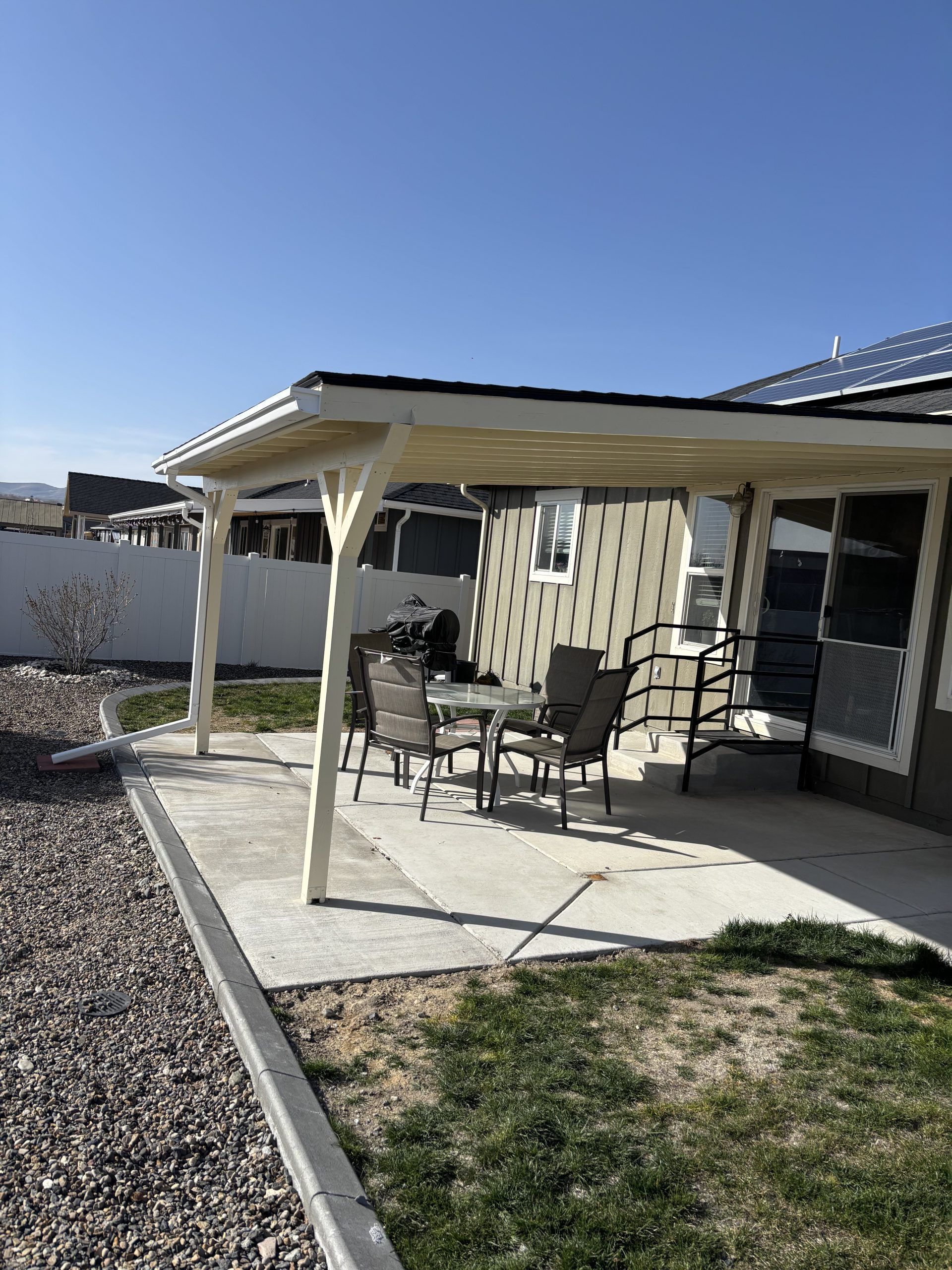 Covered patio with chairs and table beside a house, overlooking a gravel yard under a clear blue sky