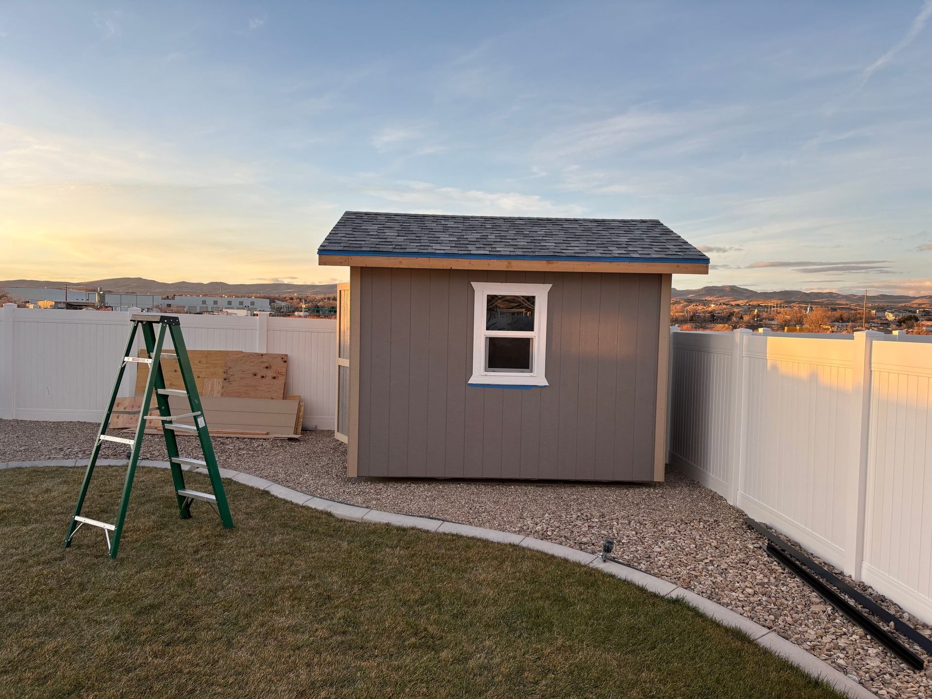 Small gray shed in a backyard at sunset, with a green ladder, gravel path, and white fence.