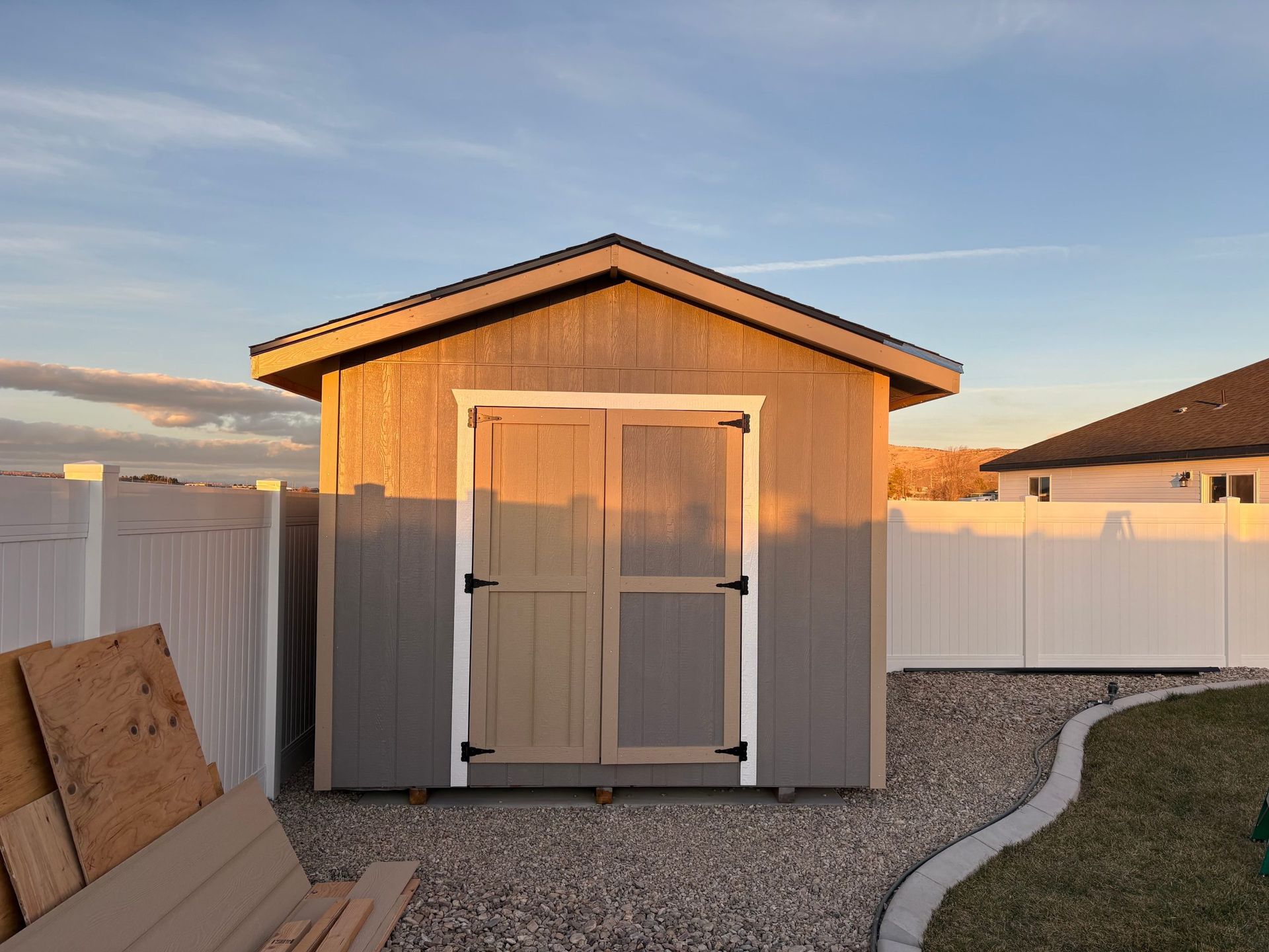 Small beige garden shed with double doors and white fencing in a gravel backyard at sunset