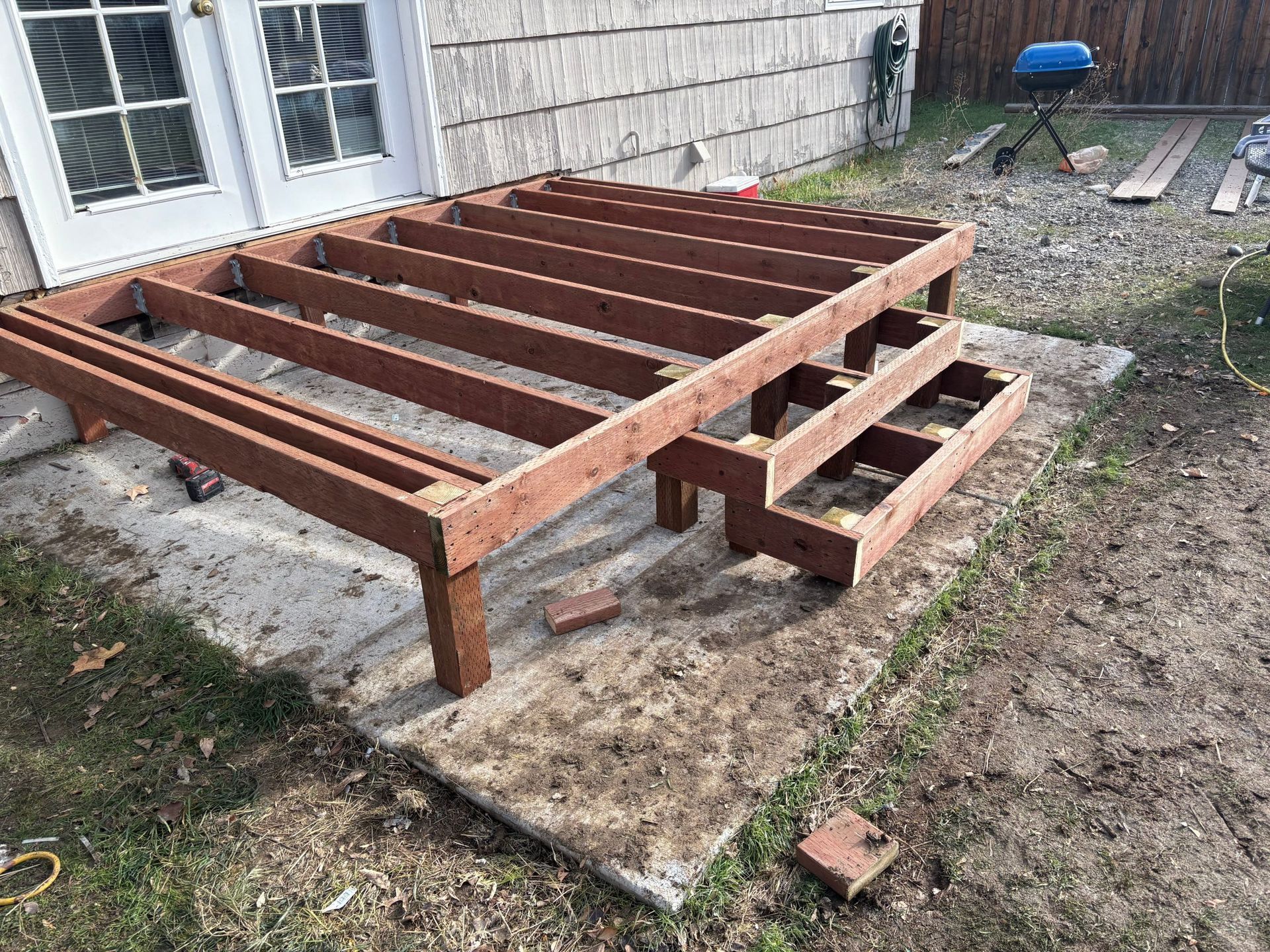 Wooden deck frame under construction beside a house, with joists on a concrete pad.