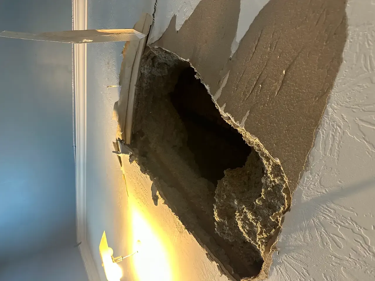 Large hole in a plaster wall with exposed insulation and broken edges, lit by sunlight.