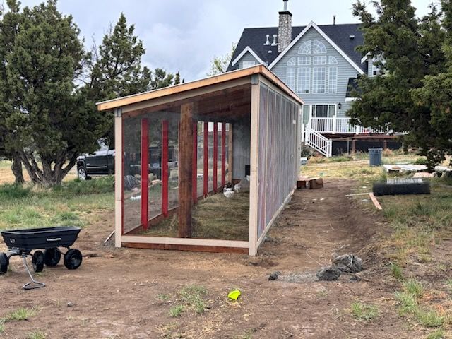 Small shed under construction in a yard, with exposed red framing and gray siding near a large house.
