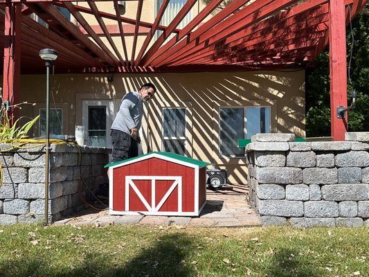 Person working by a small red-and-white shed under a red pergola, with stacked stone walls nearby.