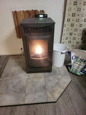 Lit pellet stove glowing in a tiled corner beside a white trash can and bag of pellets.