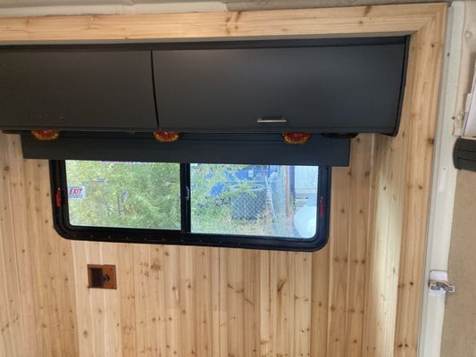 Wood-paneled RV interior with a dark window shade partially covering a window and cabinets above.