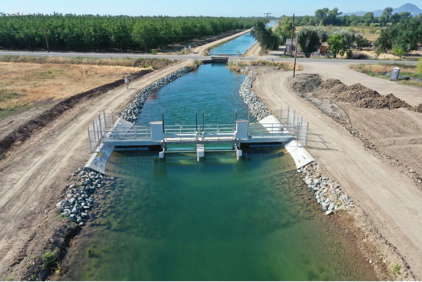Small irrigation canal with a sluice gate, lined by gravel and dirt banks in a rural field.