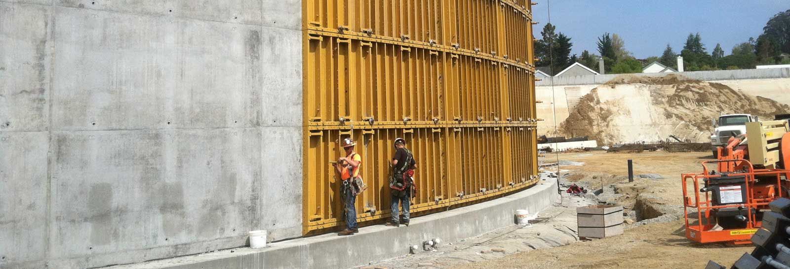 Construction site with concrete wall, yellow formwork, workers, and an excavator at the right