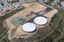 Aerial view of two large white storage tanks beside a dirt lot and residential street.