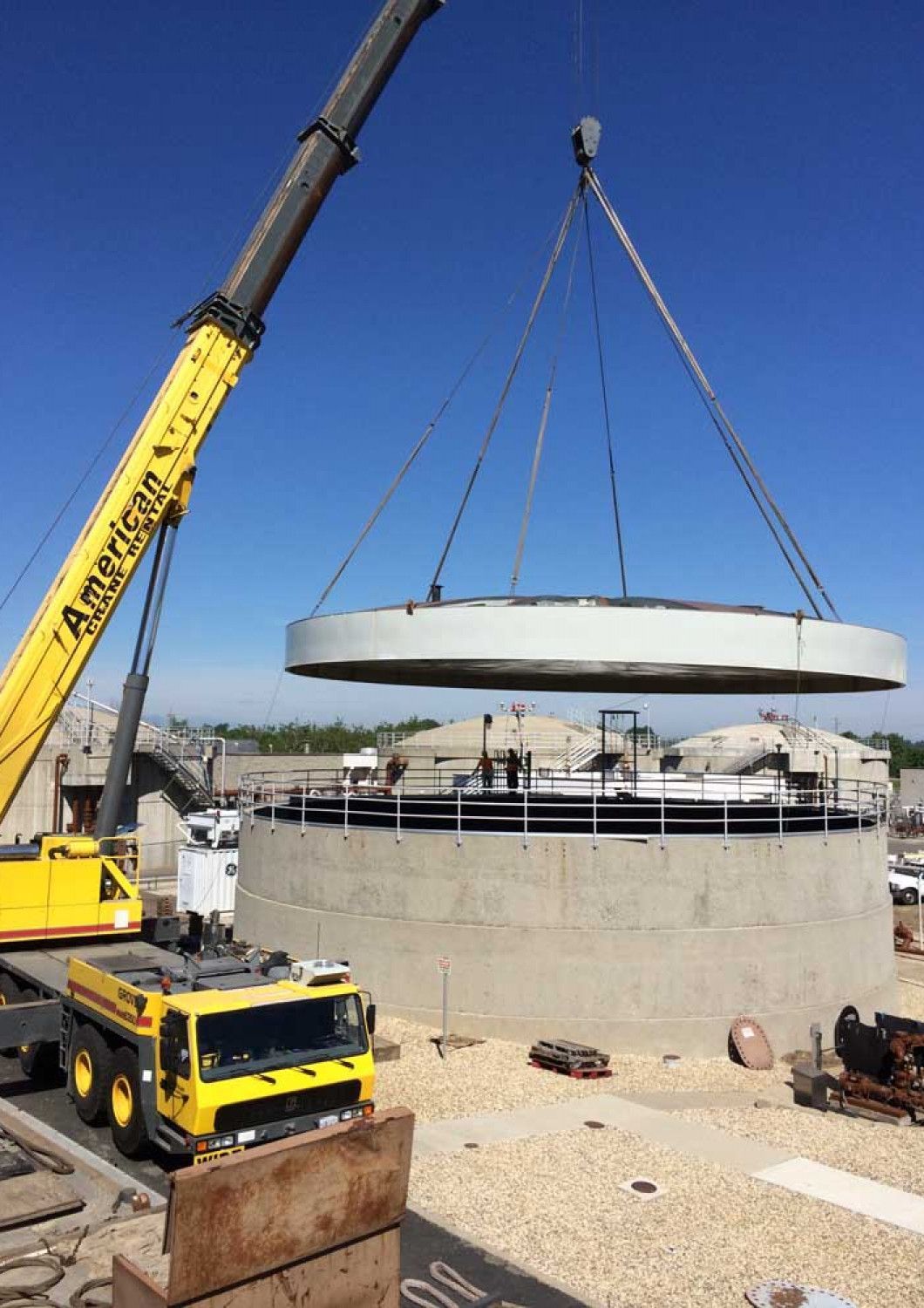 Crane lifting a large concrete ring onto a cylindrical structure at a construction site under a blue sky