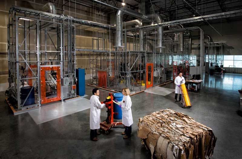 Factory workers in white coats moving wrapped goods beside industrial machinery and conveyor equipment.