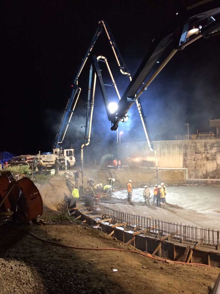 Night construction site with large cranes pouring concrete at a bridge foundation in smoky dusk light