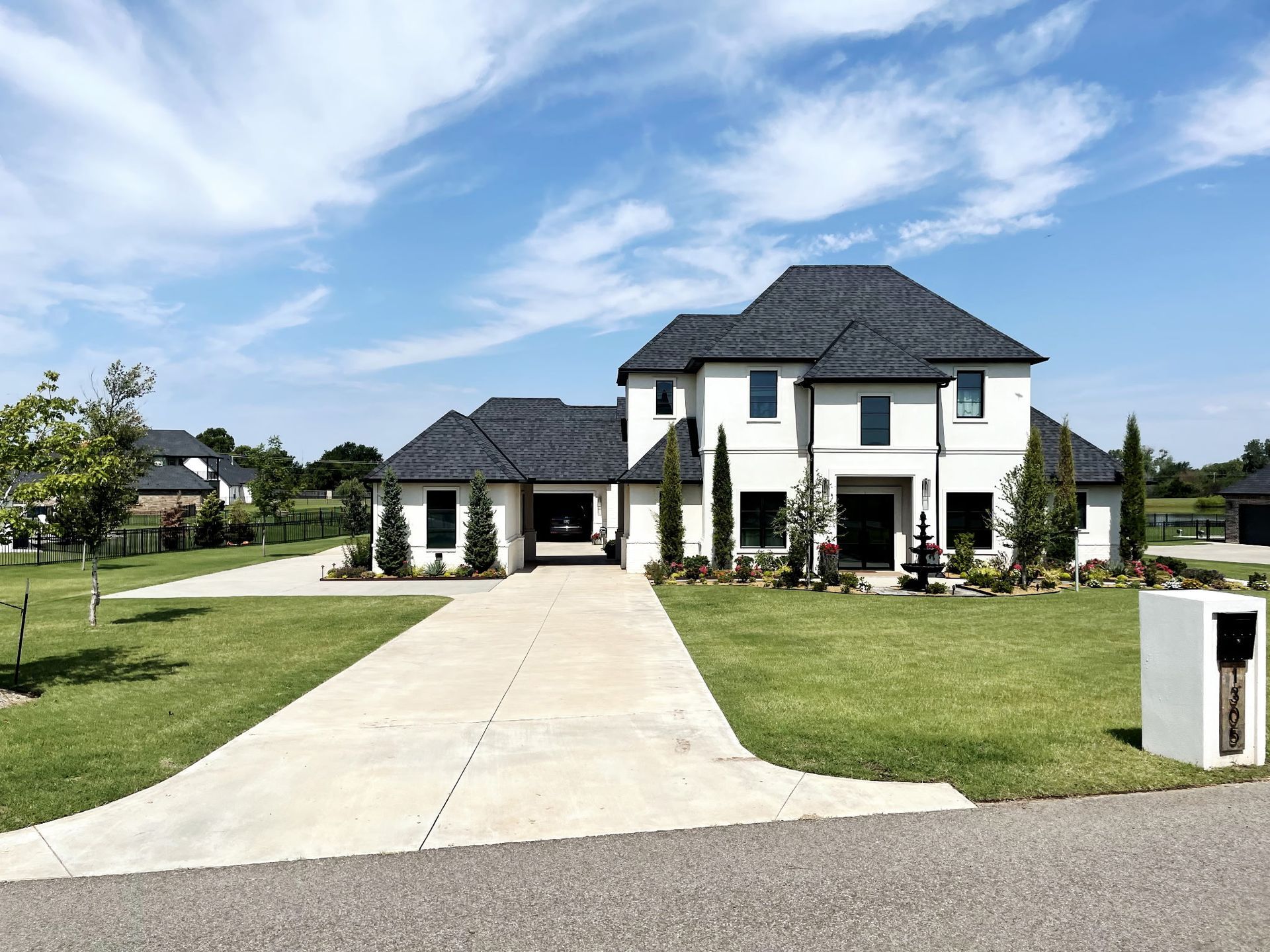 Large white house with black roof, driveway, and lush green lawn under a blue sky.