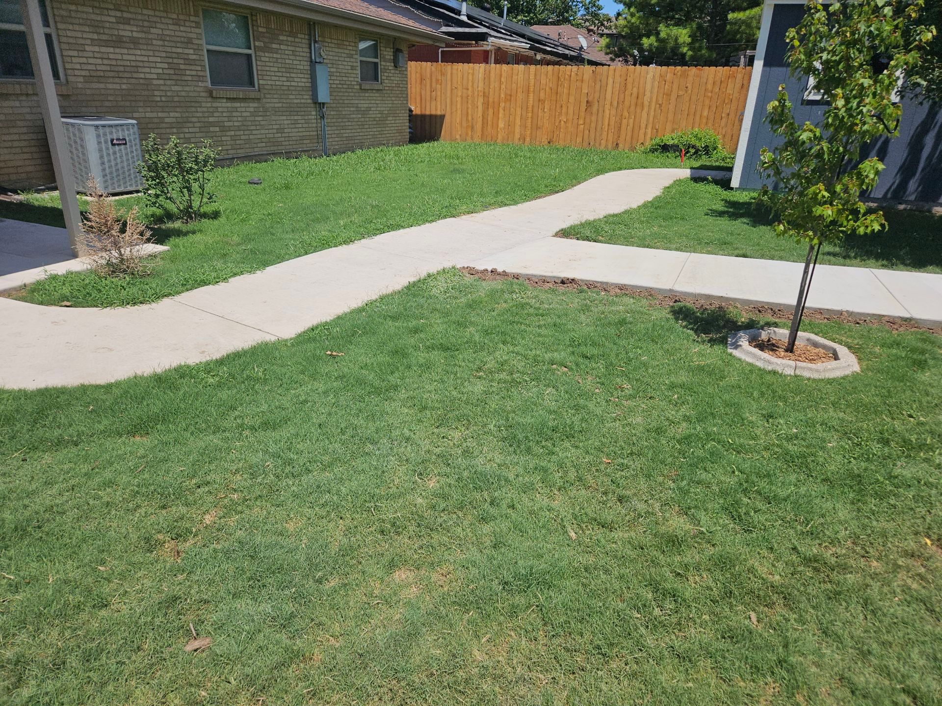 Lush green lawn with a concrete pathway leading towards a wooden fence and a house with an air conditioner.