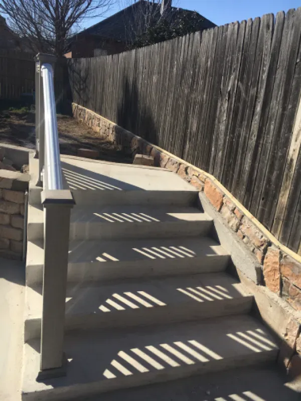 Concrete outdoor stairs with shadows, a handrail, and a stone retaining wall against a wooden fence.