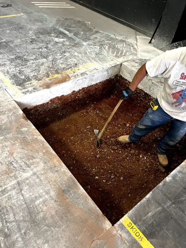 A person digs in a rectangular pit in a concrete floor, using a pickaxe.