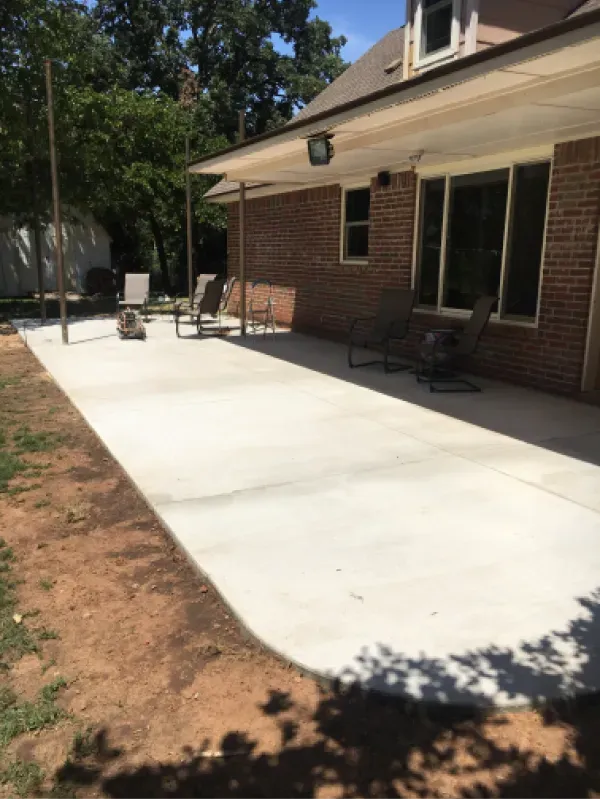 Concrete patio next to brick house with chairs, under a shaded overhang.