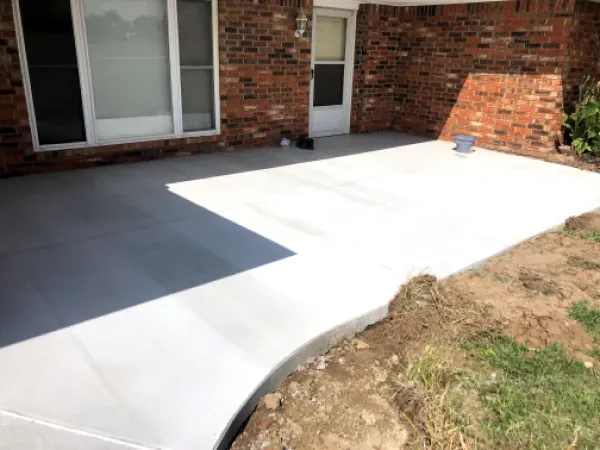 Newly poured concrete patio and walkway next to a brick building, with grass and dirt nearby.