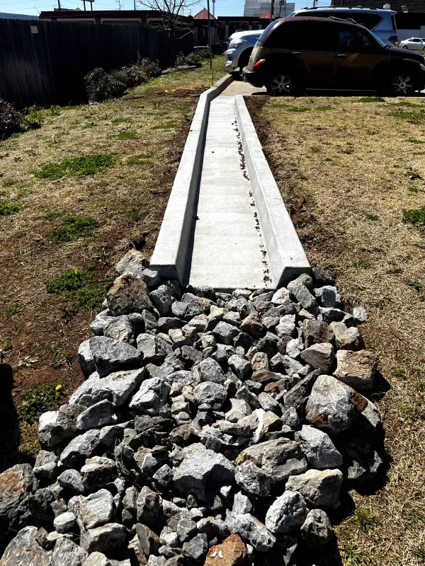 Concrete drainage channel with rock-lined approach, grass, and parked vehicles.