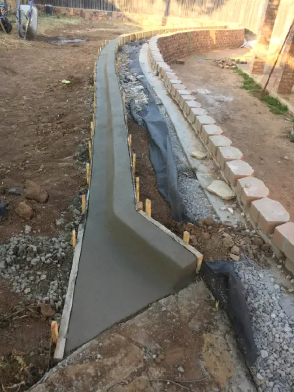 Freshly poured concrete pathway with wooden formwork, next to a rock wall in an outdoor setting.