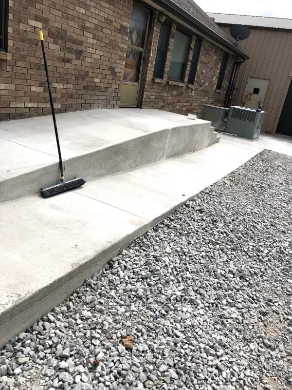 New concrete ramp and patio next to a gravel driveway, leading up to a brick building. A broom rests on the patio.