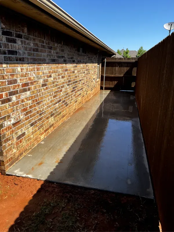 Concrete walkway between a brick wall and wooden fence, with water on the surface, under a blue sky.