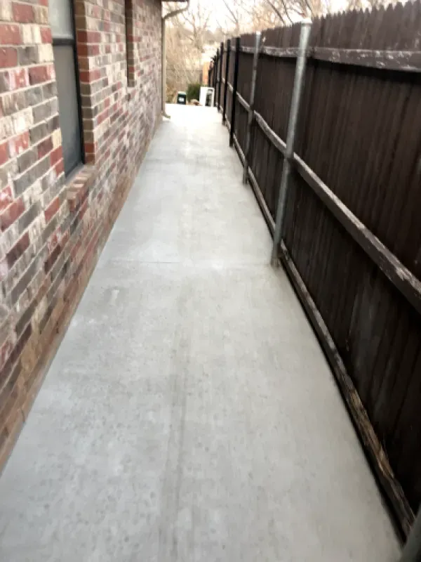 Concrete walkway between a brick building and a wooden fence.