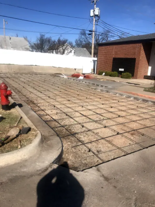 Construction site with exposed rebar grid on a prepared surface, next to a sidewalk and building.