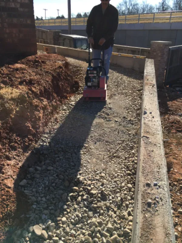 Man compacts gravel pathway with a vibratory plate compactor between concrete borders.