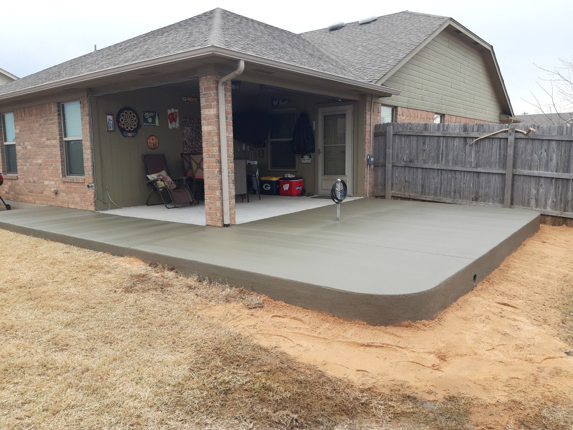 A freshly poured concrete patio next to a brick house with a covered porch and wooden fence.