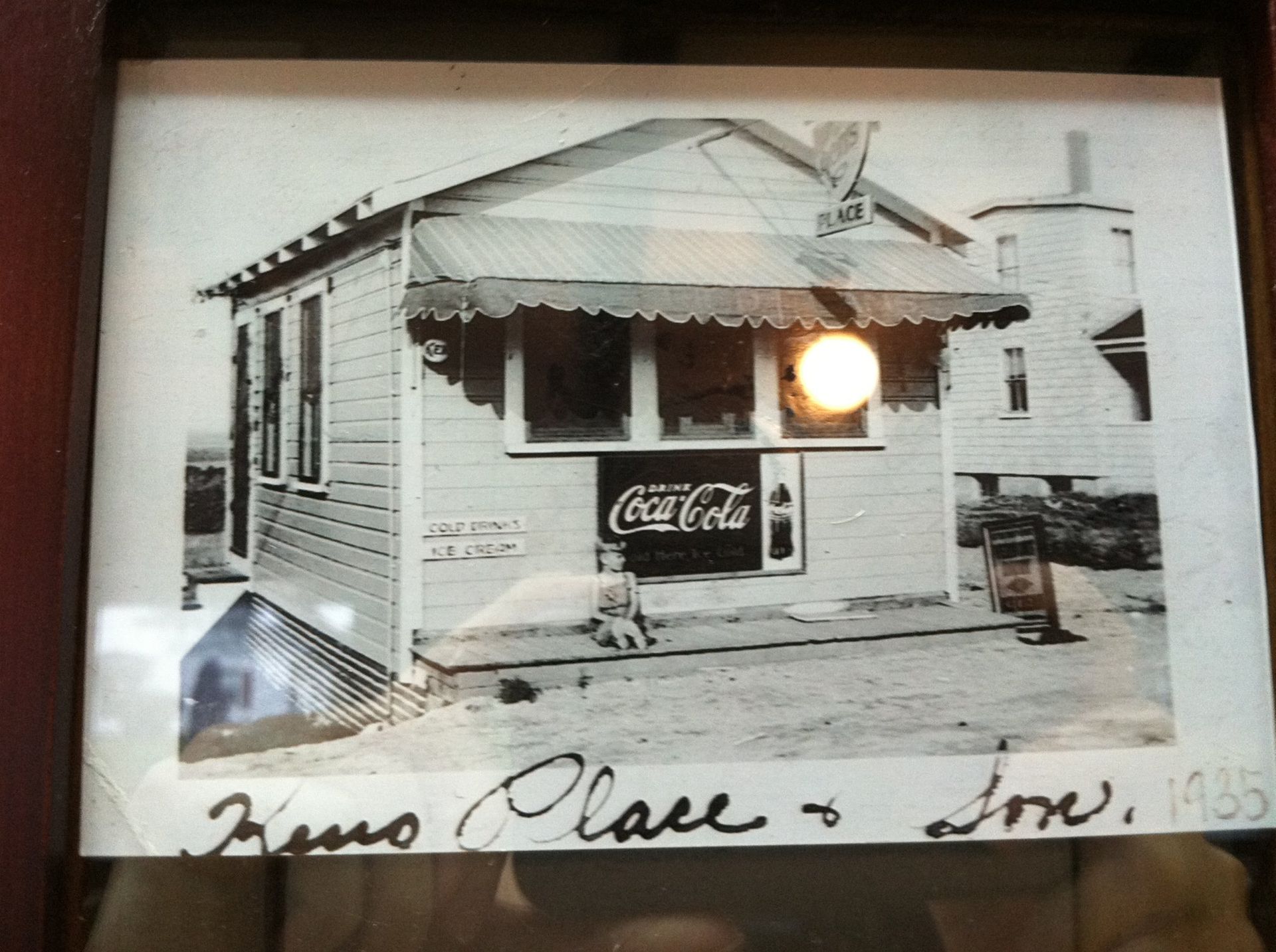 Black-and-white photo of a small wooden storefront with a Coca-Cola sign and handwritten note beneath.