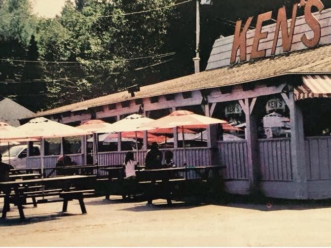 Outdoor diner patio with pink umbrellas, picnic tables, and a rustic sign reading “MEALS”