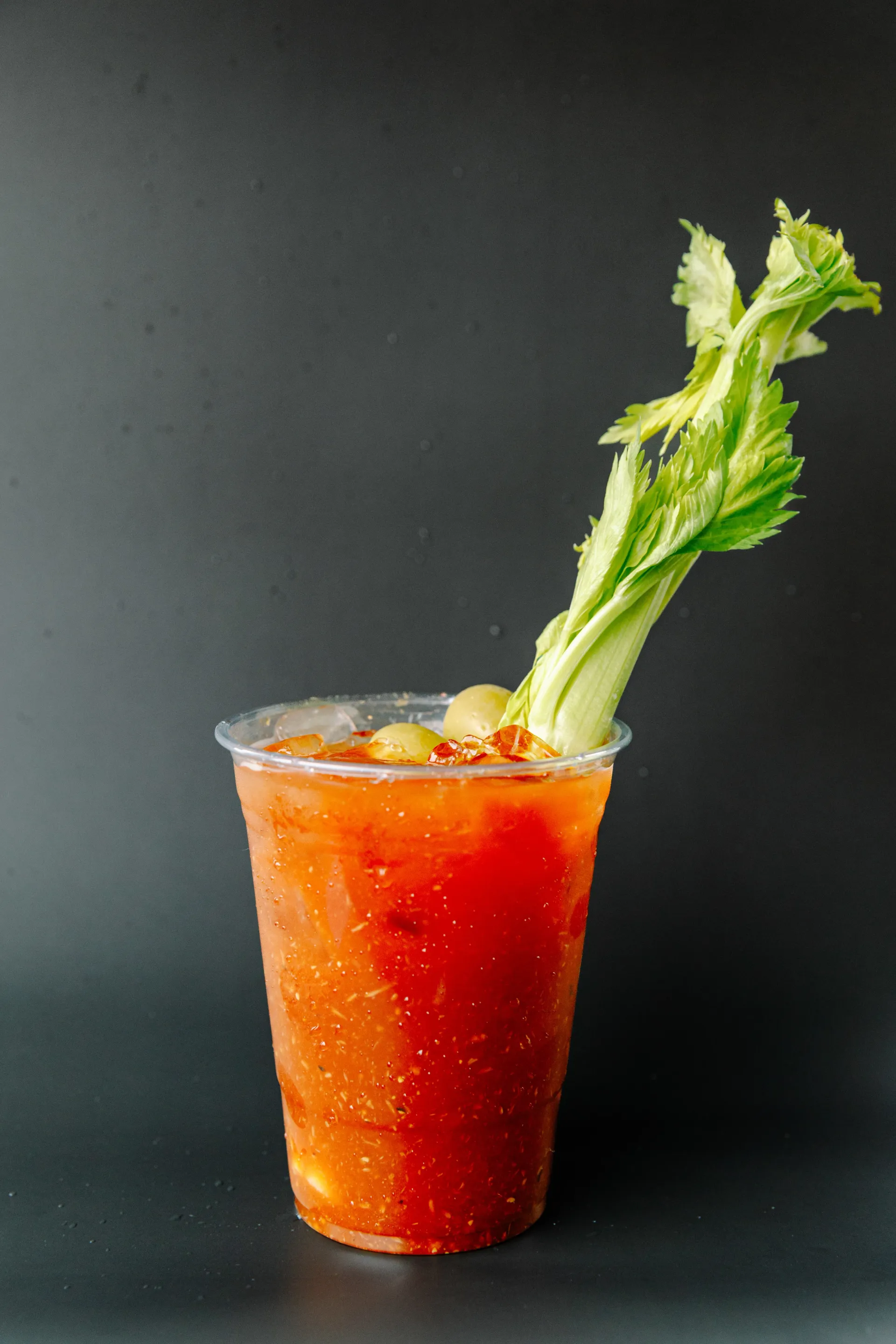 Red Bloody Mary cocktail with celery and ice in a clear cup against a dark background