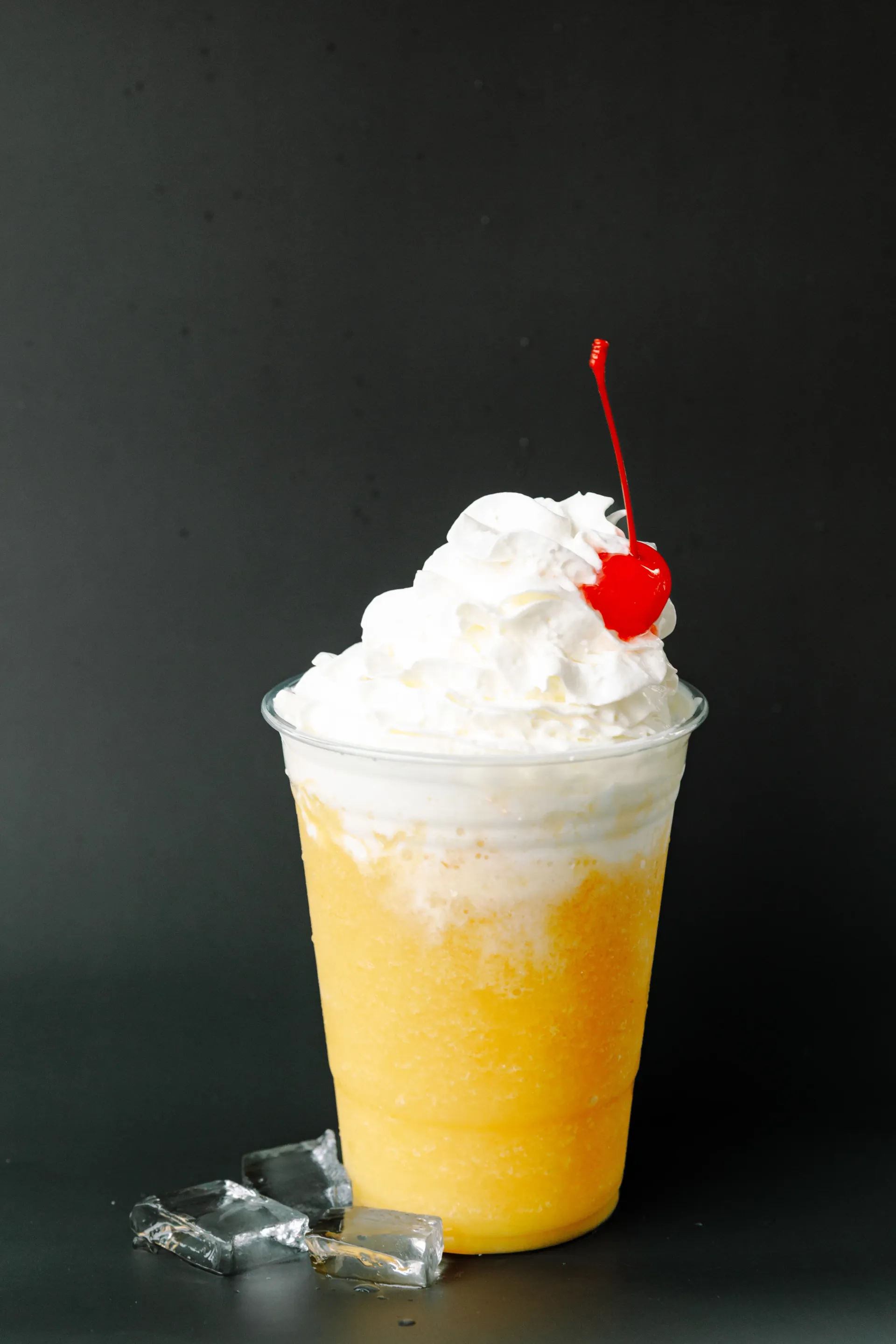 Yellow frozen drink topped with whipped cream and cherry, with ice cubes beside it on a dark background
