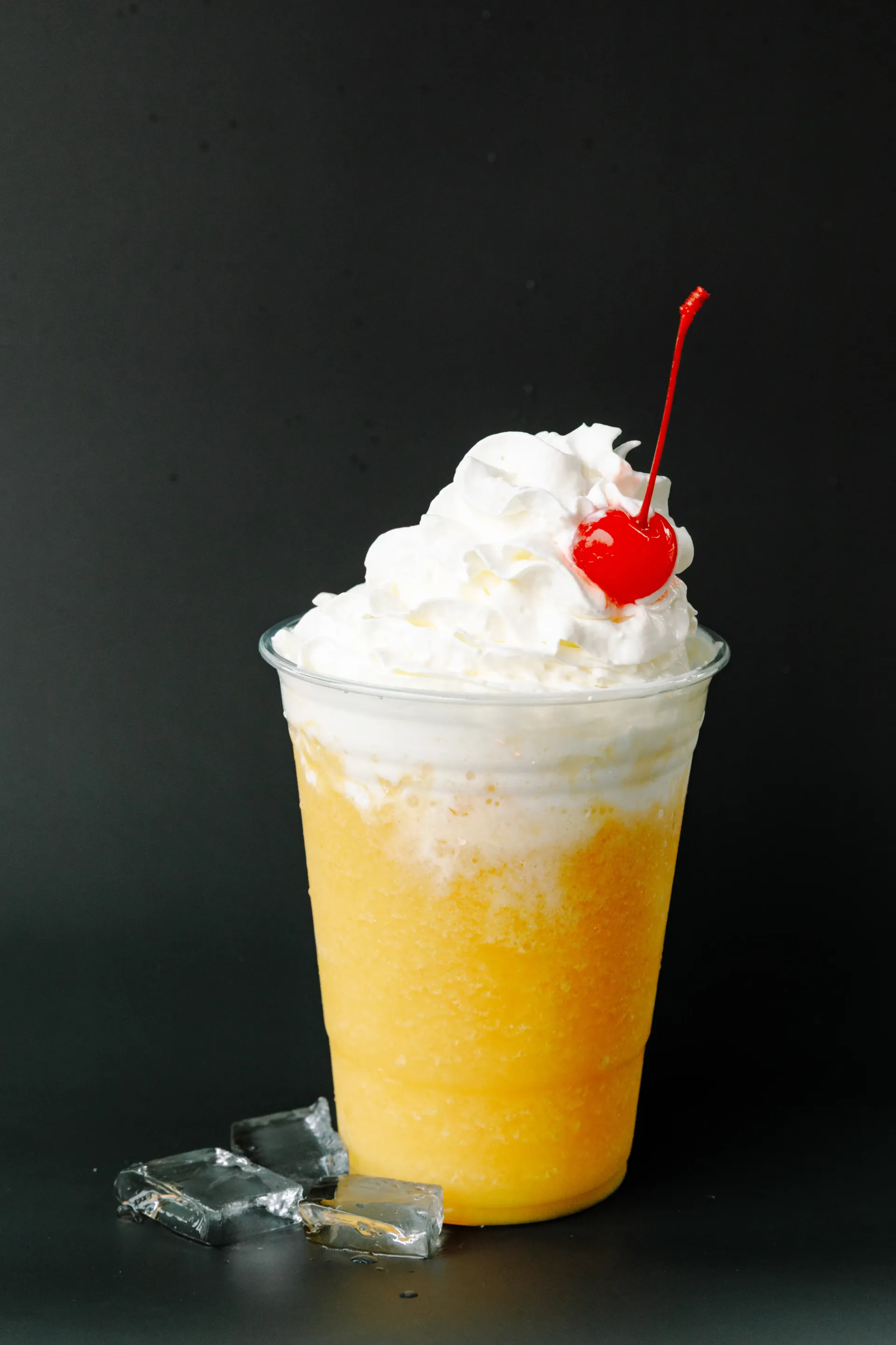 Yellow frozen drink topped with whipped cream and a cherry in a clear cup against a dark background