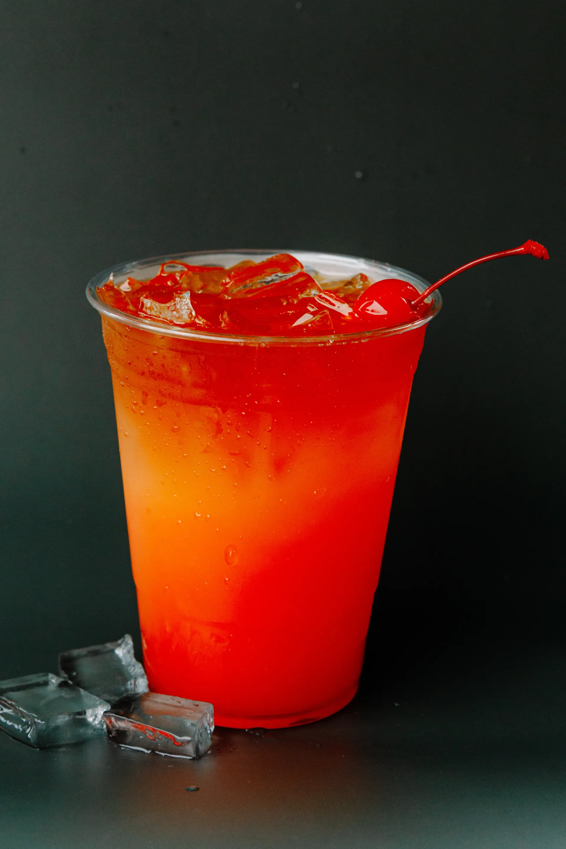 Red cocktail in a clear plastic cup with ice and a cherry garnish on a dark background