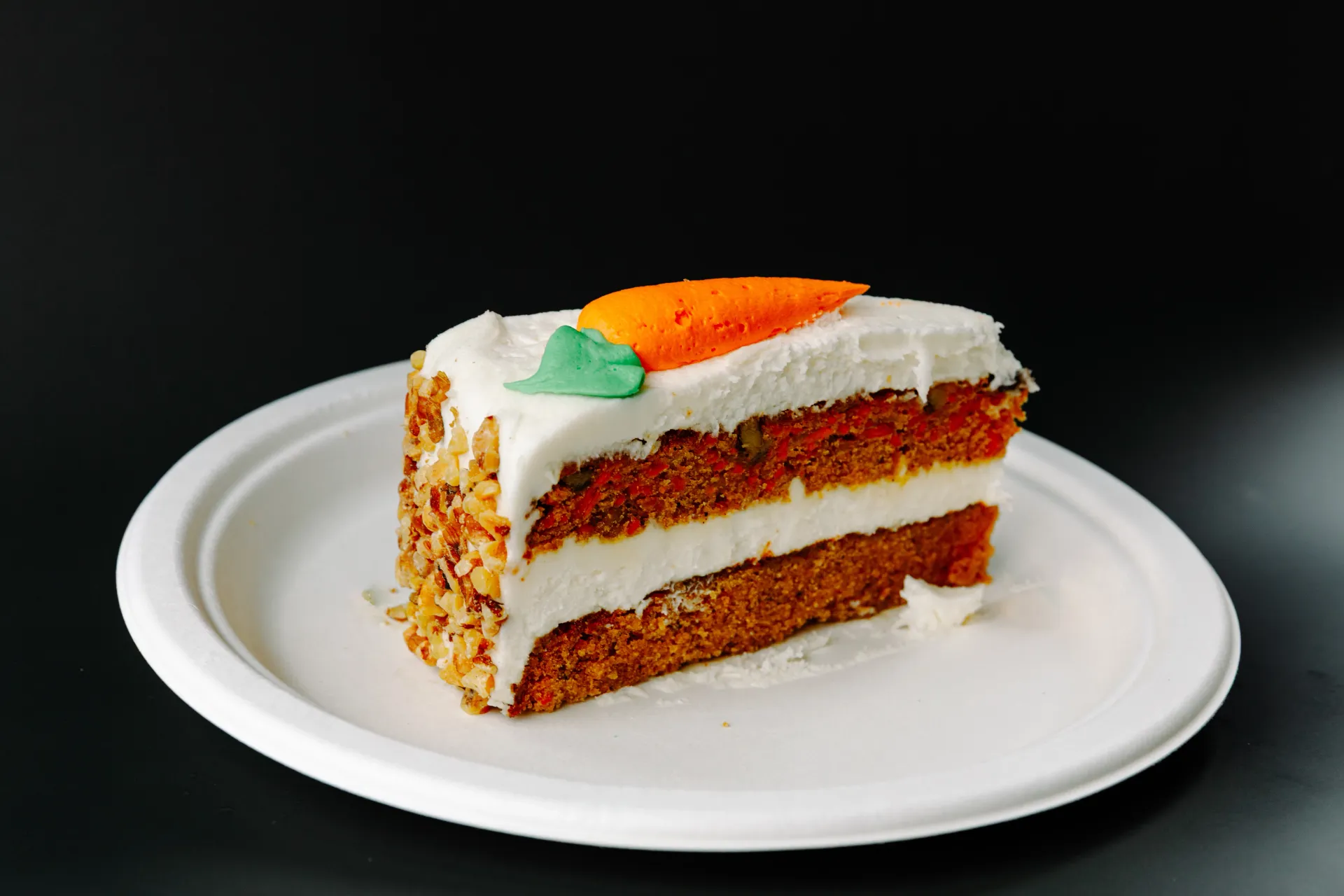 Slice of carrot cake with white frosting on a white plate against a dark background